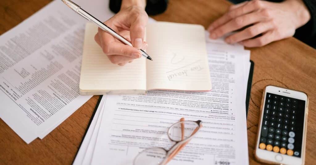 Mãos de uma pessoa fazendo anotações com documentos e calculadora sobre a mesa